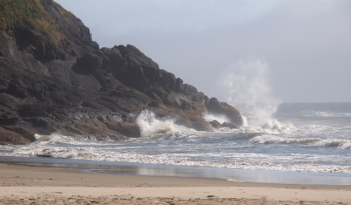 [A wave hitting rocky coast causing a spray into the air. The rest of the wave continues to the adjacent sandy shore.]